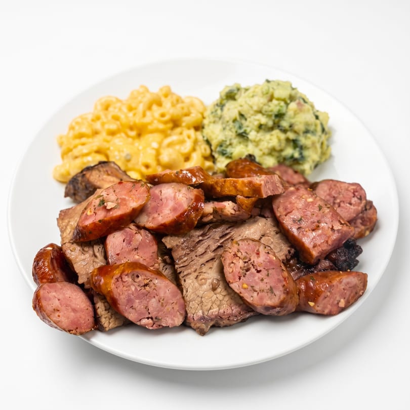 A white plate with sliced sausage, brisket, mac and cheese, and creamed spinach.