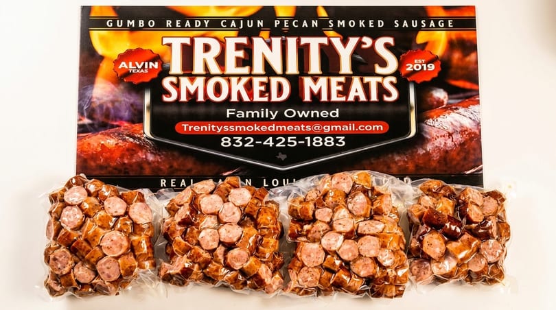 Four vacuum-sealed packages of sliced Trenity's Smoked Meats sausage in front of a brand sign.