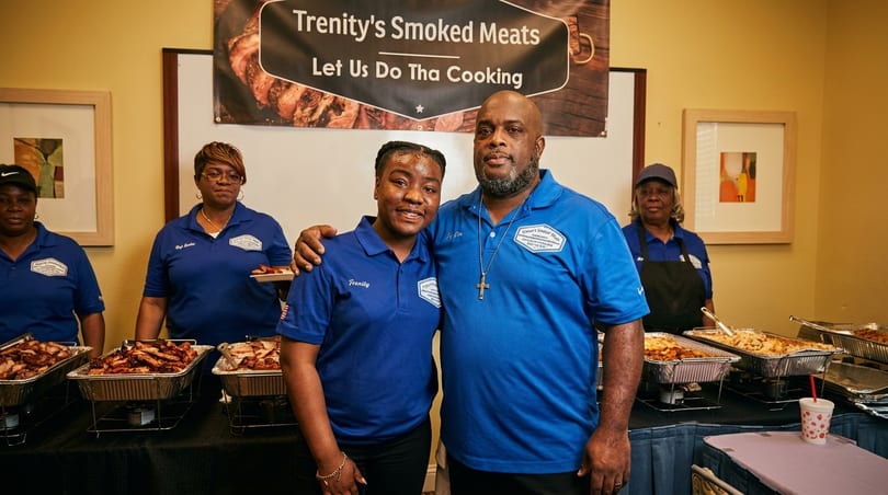 Trenity's Smoked Meats staff, including a man and woman in blue shirts, stand by buffet food.